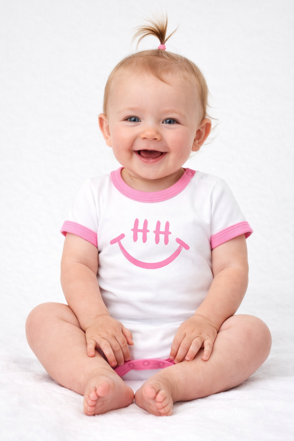 Lächelndes Baby im weißen Body mit rosa HH Smiley" Motiv, sitzt auf rosa Decke