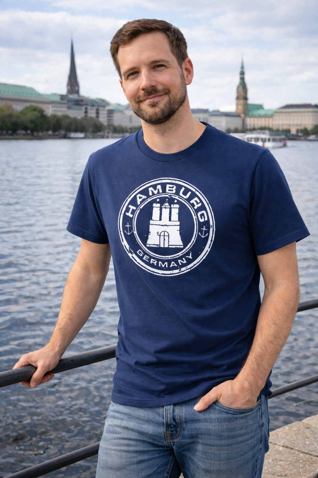 Mann steht entspannt an einem Geländer am Wasser und blickt leicht lächelnd in die Kamera. Er trägt ein dunkelblaues T-Shirt mit einem runden „Hamburg Germany“-Print, der ein stilisiertes Wahrzeichen sowie maritime Elemente zeigt. Im Hintergrund sieht man den jungfernstieg in Hamburg und die Binnenalster. Das Bild vermittelt eine ruhige, urbane Atmosphäre.
